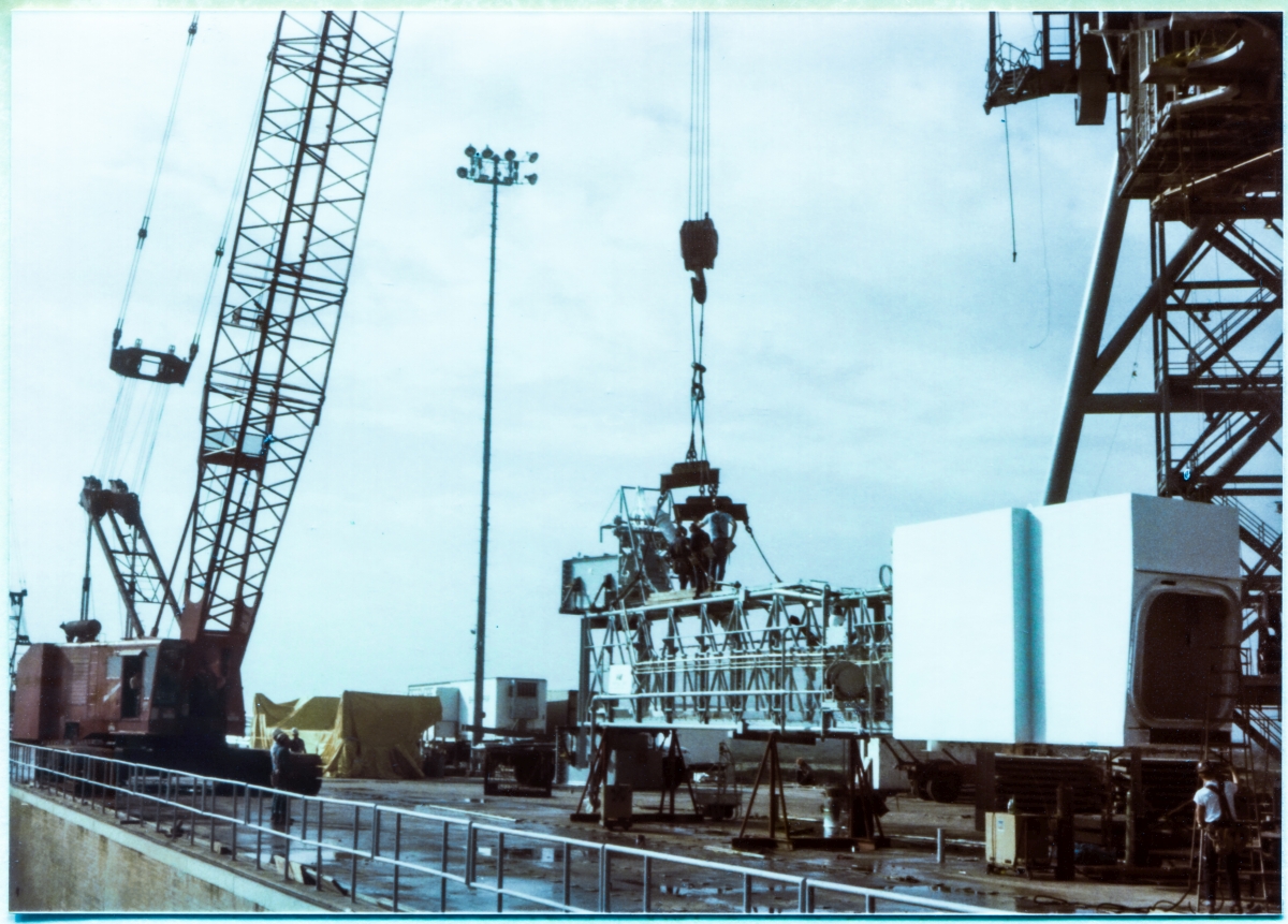 Image 130. At Space Shuttle Launch Complex 39-B, Kennedy Space Center, Florida, the Orbiter Access Arm (OAA) can be seen sitting on its support stanchions, on top of the Pad Deck, while Union Ironworkers from Local 808 working for Ivey Steel Erectors attach lifting gear to it. The crane which will lift the OAA to its final working position at Elevation 200'-0” on Side 1 of the Fixed Service Structure in a few more minutes is visible very near the west wall of the Flame Trench, in the lower left corner of the photograph. A close look at the run of handrail which guards the Flame Trench, along with the aspect of the Flame Trench Wall itself, as well as the perceived elevation of the OAA's Bottom Truss Chord, reveals that the photograph was taken from a position standing on top of one of the Spray Headers along the Flame Deflector Crest, not quite midway along its length, a bit closer to the west side of the Trench than its east side. This is a very unusual viewpoint for any image ever taken of the Pad, and was a bit hazardous to access owing to the difficulty of traversing the line of Spray Headers without getting tripped-up doing so, in conjunction with the consequences of a fall down the Fondu Fyre covered Flame Deflector Slope on either side of its Crest. Photo by James MacLaren.